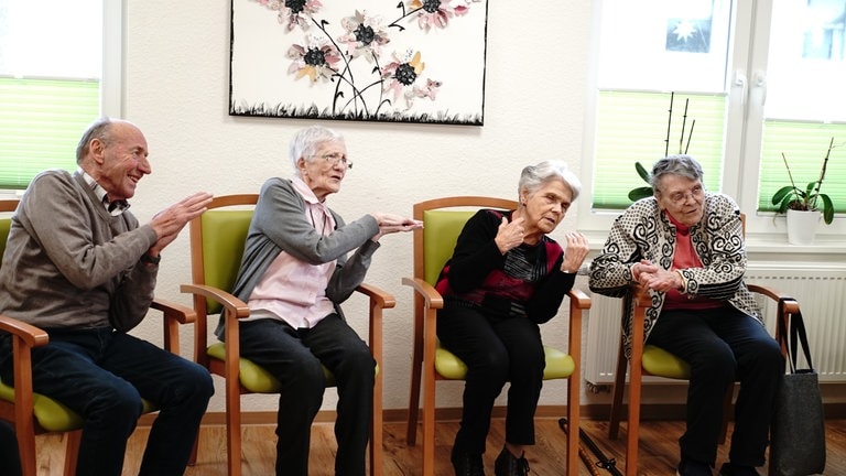Vier ältere Menschen sitzen auf Stühlen und tanzen im Sitzen. 