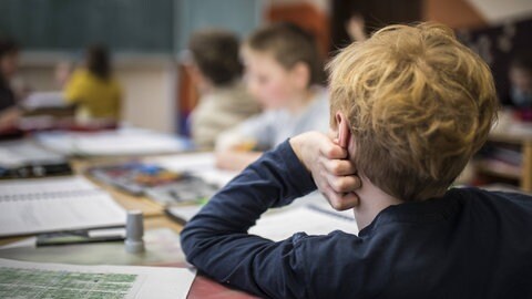 Symbolbild für ADHS und die Nachteile der Erkrankung wie sie häufig auf Social Media diskutiert werden:Junge stützt nachdenklich den Kopf in die Hand während er im Klassenzimmer sitzt und auf seine Arbeitsblätter schaut