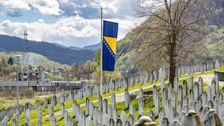 Eine bosnische Flagge weht auf Halbmast vor einer Reihe von Gräbern an der Gedenkstätte für den Völkermord von Srebrenica. Sie erinnert an die über 8.372 bosnischen Männer und Jungen, die von ihren Müttern, Schwestern und Töchtern getrennt und von bosnisch-serbischen Streitkräften Jugoslawiens getötet wurden.