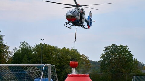 Hubschrauber nimmt Wasser auf, während eines Einsatzes gegen einen Waldbrand im Westerwald. Unter ihm ist auf einem Sportplatz ein roter Wasserbehälter, aus dem der Hubschrauber mittels einer Seilwinde neues Wasser zum Löschen auf- und mitnimmt. 