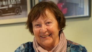 Frau sitzt an gedecktem Kaffeetisch und lächelt in die Kamera. 