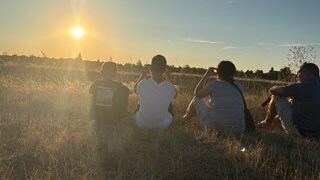 Vier Menschen sitzen mit dem Rücken zur Kamera auf einem Feld - vor ihnen die Sonne am Horizont.