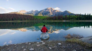 Ein Mann sitzt in einem See auf einem großen Stein und blickt auf ein Bergpanorama. In SWR1 Leute spricht Wolfgang Fasser über die große Bedeutung von Ruhe und Stille für uns.
