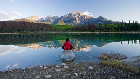 Ein Mann sitzt in einem See auf einem großen Stein und blickt auf ein Bergpanorama. In SWR1 Leute spricht Wolfgang Fasser über die große Bedeutung von Ruhe und Stille für uns.