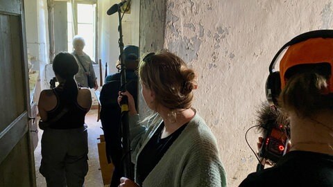 Ein Film-Team steht in einem Gang und nimmt eine ältere Frau auf. Sie erinnert sich an das SS-Massaker von 1944.