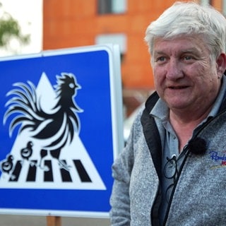 Wolfgang Rombach steht neben einem Straßenschild, auf dem Hühner auf einem Zebrastreifen zu sehen sind.