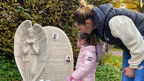Eine Frau steht mit einem kleinen Mädchen vor einem Grabstein, der mit den Bildern eines Mannes und eines Kindes geschmückt ist. An der Seite des Grabsteins ein in den Stein gemeisselter Engel im Gebet. In unserer Reihe "SWR1 Neuanfang" portraitieren wir Luana: Sie hat einen Raserunfall überlebt, ihr Mann und ihr Baby starben. Trotz des großen Traumas kämpfte sie sich zurück ins Leben.