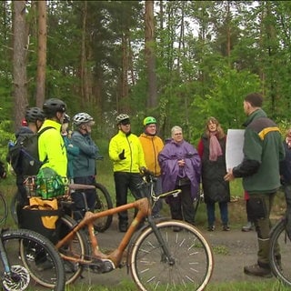 Eine Gruppe Radfahrer steht im Wald um einen Mann herum