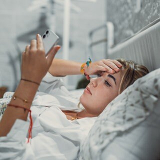 Frau liegt mit Kopfschmerzen im Bett und schaut auf ihr Handy