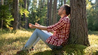 Ein Mann sitzt an einem sonnigen Tag mit geschlossenen Augen unter einer Kiefer im Wald und meditiert: Die Hälfte der Deutschen fühlt sich erschöpft. Doch Erholung braucht mehr als nur Ruhe. Sie muss zu unseren Bedürfnissen passen. 