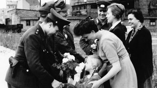 Ein Mädchen überreicht in Begleitung der Mutter Blumen an einen bewaffneten DDR-Grenzschützer. Original-Pressemeldung des DDR-Nachrichtendienstes ADN auf der Rückseite des Bildes: "Berlin 15.8.61: Berliner Bevölkerung bedankt sich bei den bewaffneten Streitkräften. Ihren Dank für den Schutz ihrer Häuser und für die gründliche Sicherung der Grenzen nach Westberlin drückten zahlreiche Berliner dadurch aus, dass sie den Streitkräften Blumen überreichten. UBz.: Am Kontrollpunkt Heinrich-Heine-Straße."