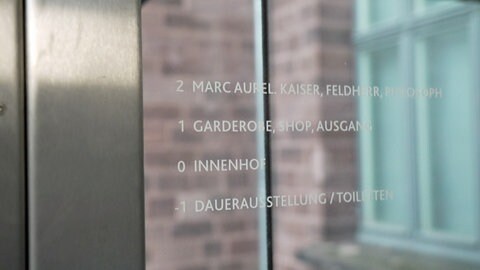 Die weiße Schrift im Glasaufzug ist klein und hoch platziert. Menschen im Rollstuhl oder mit eingeschränktem Sehvermögen haben Mühe, sie zu erkennen.