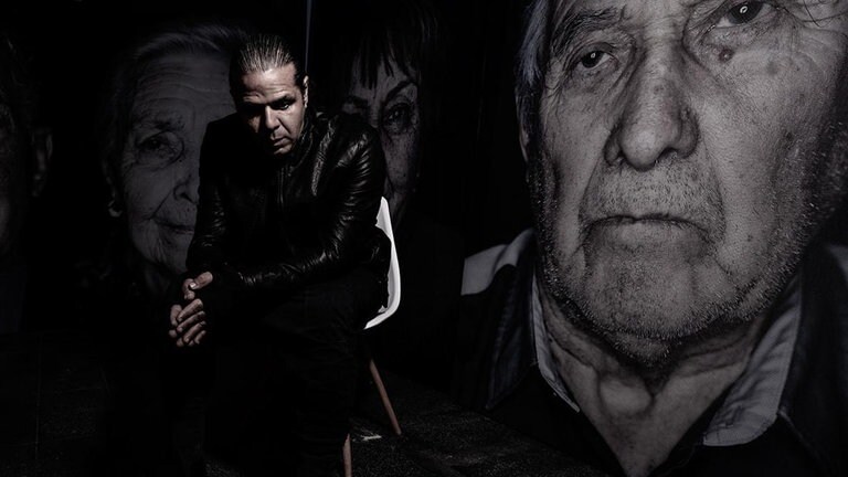 Fotograf Luigi Toscano sitzt auf einem Stuhl, dahinter drei Portaits von Holocaust-Überlebenden.
