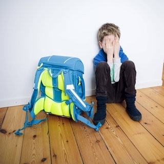 Symbolfoto zum Thema Angst vor der Schule. Ein Junge sitzt traurig in seinem Kinderzimmer neben seinem Schulranzen und versteckt sein Gesicht hinter seinen Haenden.