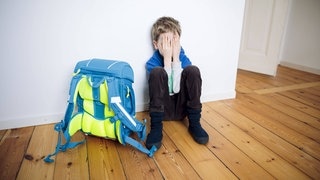 Symbolfoto zum Thema Angst vor der Schule. Ein Junge sitzt traurig in seinem Kinderzimmer neben seinem Schulranzen und versteckt sein Gesicht hinter seinen Haenden.