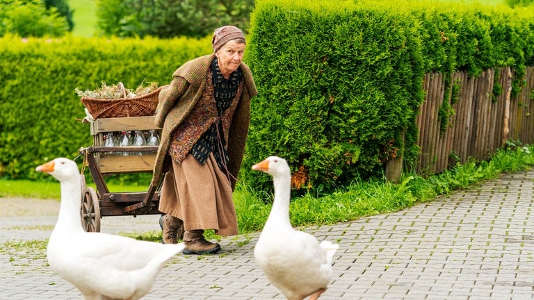 Lioba (Lisbeth Felder) zieht ihren Leiterwagen hinter sich her, im Vordergrund zwei Gänse