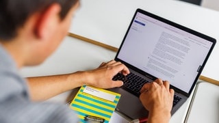 Ein Schüler eines Gymnasiums sitzt vor einem Laptop und benutzt ein KI-Tool, vor ihm eine Lektürehilfe zur Georg Büchners "Woyzeck": KI verändert das Lernen in der Schule. 