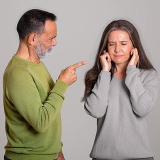 Wütender Mann zeigt mit Finger auf Frau; die hält sich die Ohren zu: Emotionale Gewalt ist oft schwer zu erkennen. Die Folgen sind jedoch gravierend. Gleichzeitig gilt: Nicht jedes böse Wort ist ein Übergriff. 