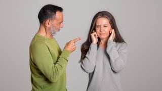 Wütender Mann zeigt mit Finger auf Frau; die hält sich die Ohren zu: Emotionale Gewalt ist oft schwer zu erkennen. Die Folgen sind jedoch gravierend. Gleichzeitig gilt: Nicht jedes böse Wort ist ein Übergriff. 