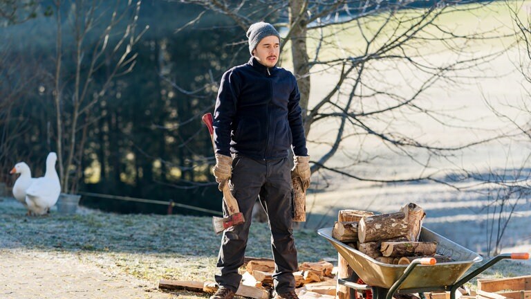 Sebastian (Dominik Stricker) steht mit der Axt in der Hand vor einer Schubkarre, die mit Holz beladen ist