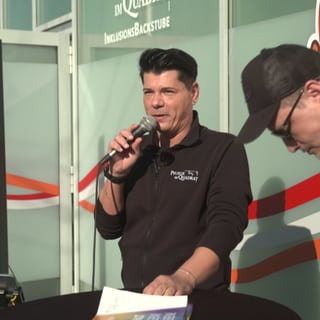 Panajotis Neuert steht an einem Stehtisch und hält ein Mikrofon in seiner Hand. 