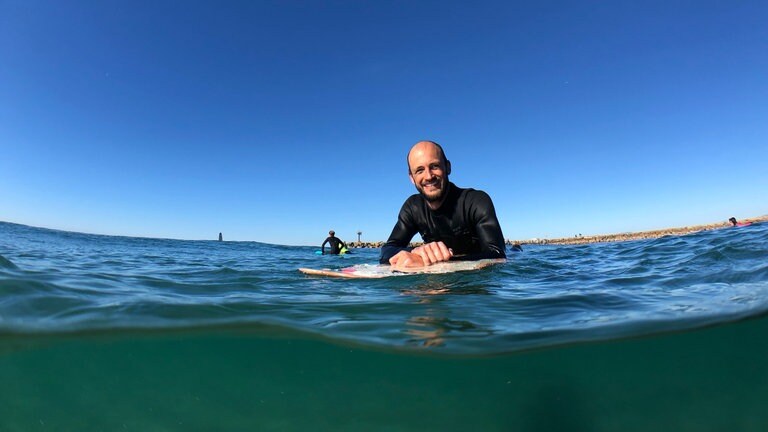 Der querschnittgelähmte Johannes Laing ist im Wasser, stützt sich auf sein Surfbrett und grinst in die Kamera.