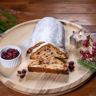 Ein nach SWR4 Rezept fertig gebackener Stollen mit Cranberry und Marzipan angeschnitten auf einem runden Holzbrett mit einer brennenden Kerze in Sternform und einer Schale getrockneter Cranberrys