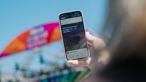 Ein Handy mit der Landingpage swr.desommerfestival vor der Bühne des SWR auf dem Stuttgarter Schlossplatz