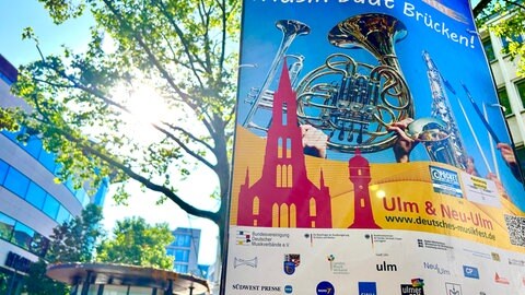 Ein Plakat zum Deutschen Musikfest im Vordergrund. Dahinter drängt sich die Sonne durch die Äste eines Baums. 