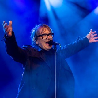 Sven Regener steht vor einem Mikrofon und reißt die Arme hoch: Der Schriftsteller und Musiker wird der Mainzer Stadtschreiber 2026