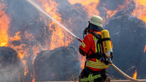 Ein Feuerwehrmann löscht brennende Ballen. (Symbolbild) Der Stadtrat in Ingelheim hat entschieden, dass mehr hauptamtliche Feuerwehrleute die Ehrenamtler entlasten sollen. 