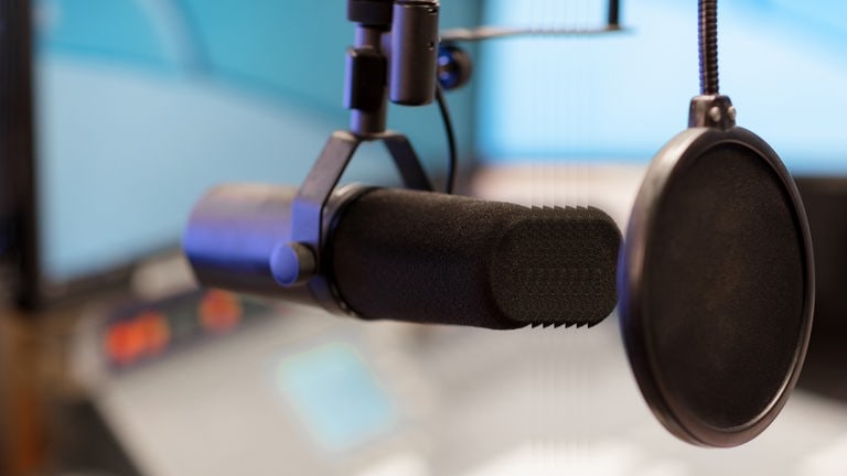 Ein Mikrofon mit Ploppschutz in einem Studio, im Hintergrund ein Mischpult mit Monitor