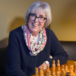 Als ihr Mann 1975 vier Wochen in der Kur ist, lernt Hanne das Schachspielen. Seitdem hat sie der Sport nicht mehr losgelassen. Hanne sitzt an einem Tisch vor dem Schachbrett. Sie lacht in die Kamera.