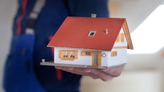 Symbolbild: Ein Handwerker hält ein Modell von einem Haus. | Die eigene Wohnung oder das eigene Haus kann durch einen Umbau altersgerecht werden.