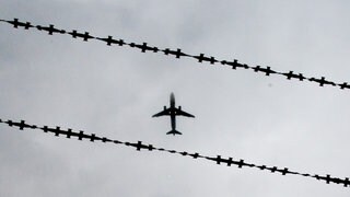 Ein Flugzeug am Himmel, durch Stacheldraht fotografiert. Ein aggressiver Flüchtling aus Windesheim im Kreis Bad Kreuznach soll mit so einem Flieger abgeschoben werden (Symbolbild)