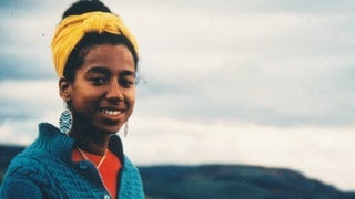 Die Schriftstellerin May Ayim: Eine junge schwarze Frau lächelt in die Kamera, ihre Haare sind nach oben gebunden, sie trägt ein gelbes Haarband, große Ohrhänger, einen blauen Strickpullover. Im V-Ausschnitt des Strickpullover sieht man ein rotes T-Shirt und eine goldene Kette. Im Hintergrund ist hügelig-waldige Natur unscharf zu erkennen, der Himmel ist gräulich-bewölkt.