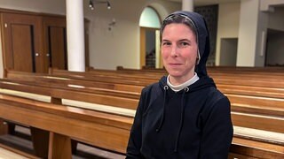 Schwester Marie-Salome sitzt auf einer Kirchenbank und lächelt in die Kamera. Sie trägt eine schwarze Haube, die für Schwestern typische Kleidung. 