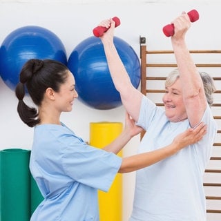 Seniorin wird beim Hanteltraining von einer Therapeuting unterstützt: Wie körperlich gesund und psychisch stabil ein Mensch vor einer OP oder Krebstherapie ist, ist mitentscheidend für den Behandlungserfolg. Deshalb bieten einige Kliniken ihren Patient*innen eine mehrwöchige "Prähabilitation" an. In dieser Zeit bauen sie Muskeln auf, lernen tiefer zu atmen und sich zu entspannen, ernähren sich gesünder, machen Kognitionsübungen. 