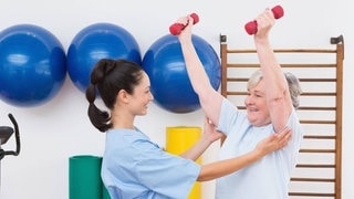 Seniorin wird beim Hanteltraining von einer Therapeuting unterstützt: Wie körperlich gesund und psychisch stabil ein Mensch vor einer OP oder Krebstherapie ist, ist mitentscheidend für den Behandlungserfolg. Deshalb bieten einige Kliniken ihren Patient*innen eine mehrwöchige "Prähabilitation" an. In dieser Zeit bauen sie Muskeln auf, lernen tiefer zu atmen und sich zu entspannen, ernähren sich gesünder, machen Kognitionsübungen. 