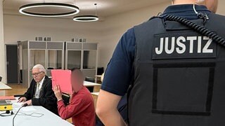 Ein Anwalt sitz in schwarzer Robe neben dem Angeklagten, der sich eine rote Mappe vor das Gesicht hält. Im Vordergrund rechts sieht man den Rücken eines Justizbeamten.