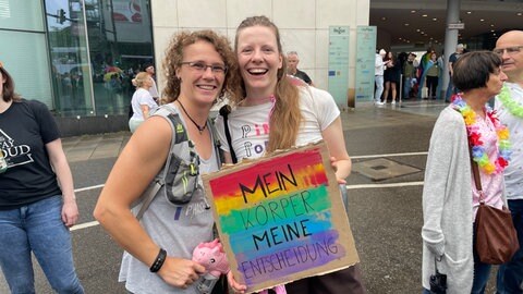 Sabrina (links) und Frederike (rechts) schauen beim CSD Stuttgart in die Kamera und ein Schild mit der Aufschrift "Mein Körper meine Entscheidung"
