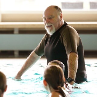 Ein Mann mit weißem Bart und Glatze ist mit drei Kindern im Schwimmbecken