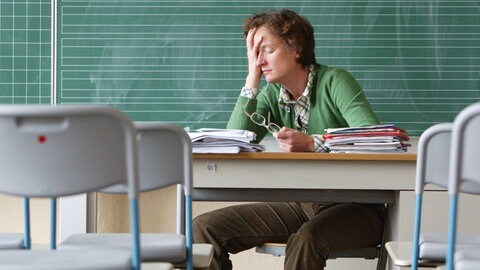 Lehrerin sitzt alleine im Klassenzimmer und stützt erschöpft den Kopf in die Hand: In ganz Deutschland geben quer durch alle Schulformen etliche Lehrkräfte ihren Beruf auf, obwohl sie ihn lieben. Die Gründe sind vielfältig: zu wenig Zeit für die Kinder, kaum individuelle Förderung, zu viel Stress.