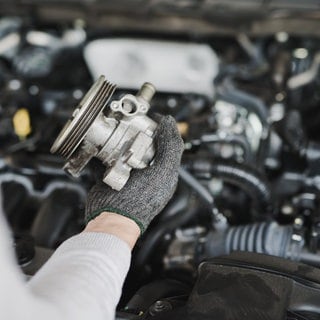 Zu sehen ist die Hand einer Person, die vor einem Auto mit geöffneter Motorhaube steht und ein Autoersatzteil hält.