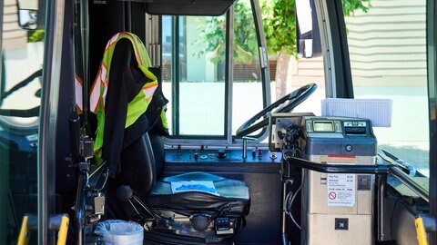 Detailaufnahme des Fahrerarbeitsplatzes in einem städtischen Bus, inklusive Steuerungspanel und Fahrersitz. Die Warnweste und das Ticketing-System zeigen die Alltagsausstattung im öffentlichen Nahverkehr.
