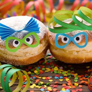 SWR1 Pfännle meets Fasnacht - wir feiern Fasnet, Fastnacht, Fasching und Karneval kulinarisch! Küchenkäpsele und Rezepte für Fasnachtsküchle, Nonnenfürzle und mehr.  Das Bild zeigt einen Berliner  Krapfen im Faschingskostüm: Maske, Konfetti und Luftschlangen