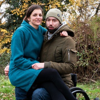 Eine lächelnde Frau sitzt auf dem Schoß ihres Mannes, der im Rollstuhl sitzt
