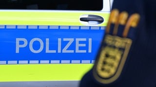 Ein Polizist vor einem Polizeiauto (Symbolbild). Die Polizei sucht mit Hochdruck nach einem weißen Kleintransporter. Der Fahrer könnte für den tödlichen Unfall auf der A3 bei Wertheim verantwortlich sein. 