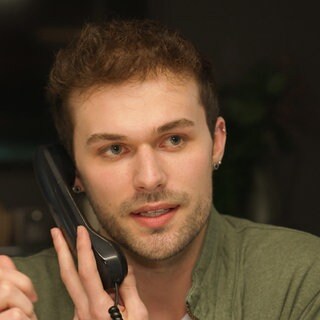Azubi Kevin sitzt in einem dunklen Raum und hält einen Telefonhörer an sein Ohr.
