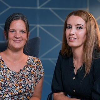 Karin und Peri leben mit ihren Familien zusammen im Hoffnungshaus. Sie sitzen vor einer dunkelblauen Wand mit glänzenden Mustern, nebeneinander auf zwei Sesseln. Karin hat ihre Haare in einem Pferdeschwanz und trägt eine geblümte Bluse. Peri trägt ihre langen, braunen Haare offen und ein schwarzes Sommerkleid. 